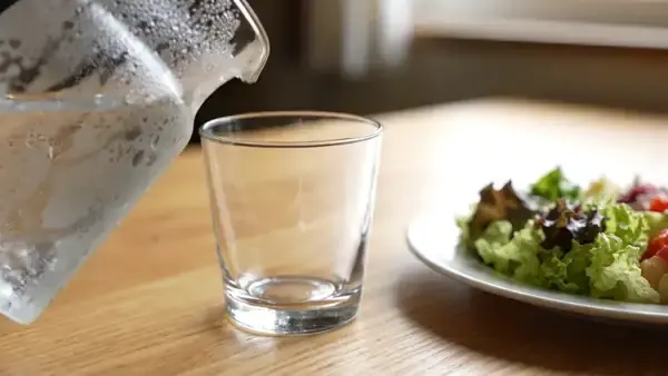 Cold water pouring from a clear pitcher into a short glass beside a lunch plate